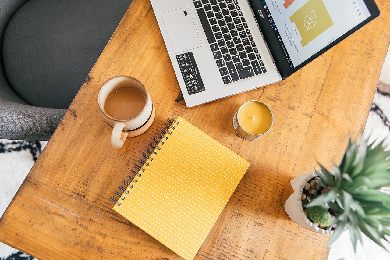 An overhead shot of Rachel’s desk. The desk is wooden; on it sits a laptop, and on the screen is a page from The Ethical Copywriter website. There is also a yellow notebook, yellow candle, cup of coffee and plant on the desk. 