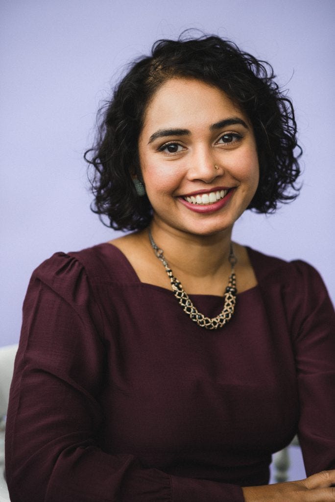 Photography of SJ Sindu, a brown woman with chin length curly black hair. She smiles broadly at the camera and is wearing a thick gold and black necklace and a burgandy top