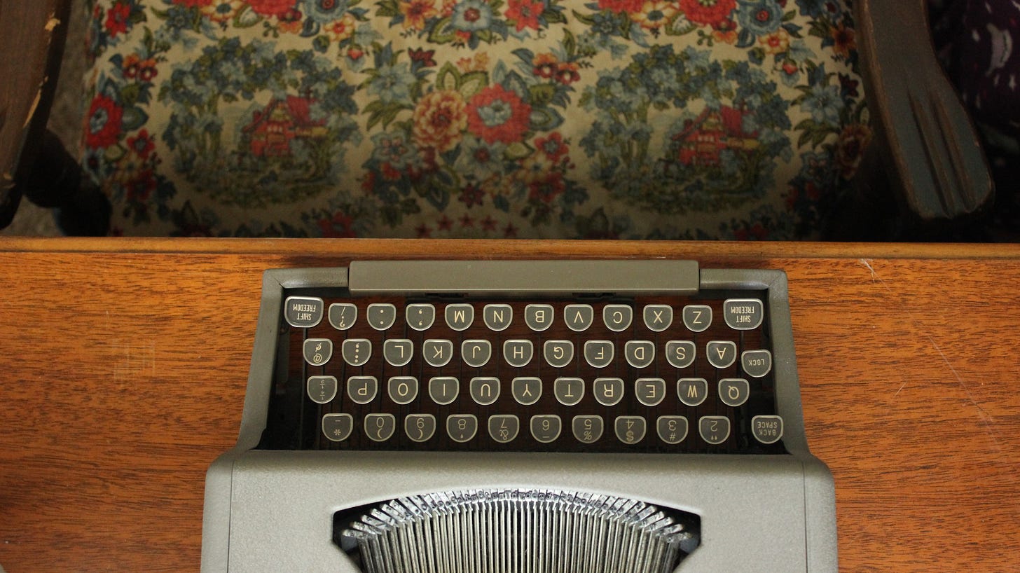 bird's eye view of a manual typewriter on a scuffed wooden desk; part of a chair with floral upholstery can be seen at the top of the image