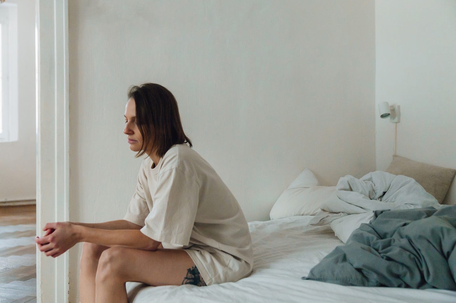 A woman in an oversized t-shirt sits sadly on the edge of a rumpled bed.