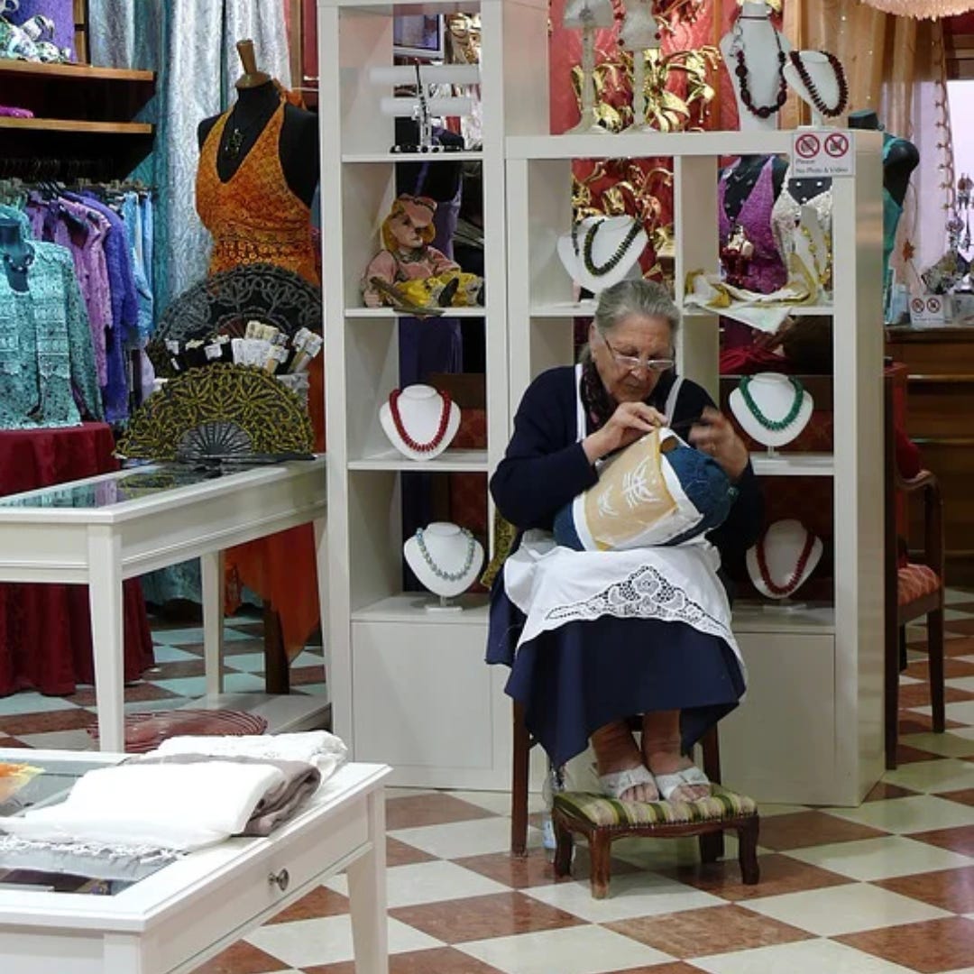  lacemaker at work. A grandmotherly elder women sits in the center of clothing store and stitches her lace work. She wears a classic white apron with a lace border and is surrounding by many textiles - fans, dresses and also jewelry. 