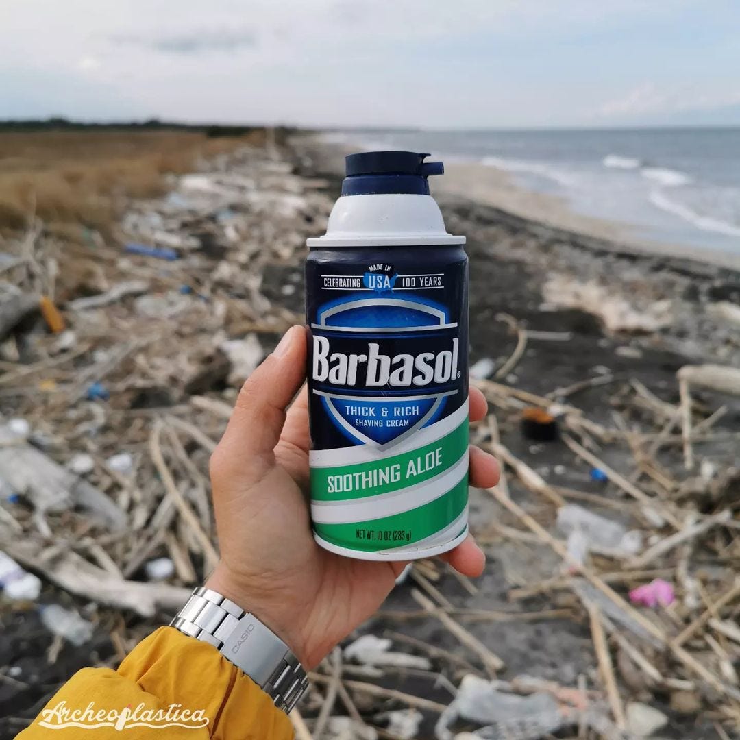 Una mano afferra una confezione di Barbasol, un prodotto fatto di aloe; sullo sfondo, una spiaggia piena di rifiuti.
