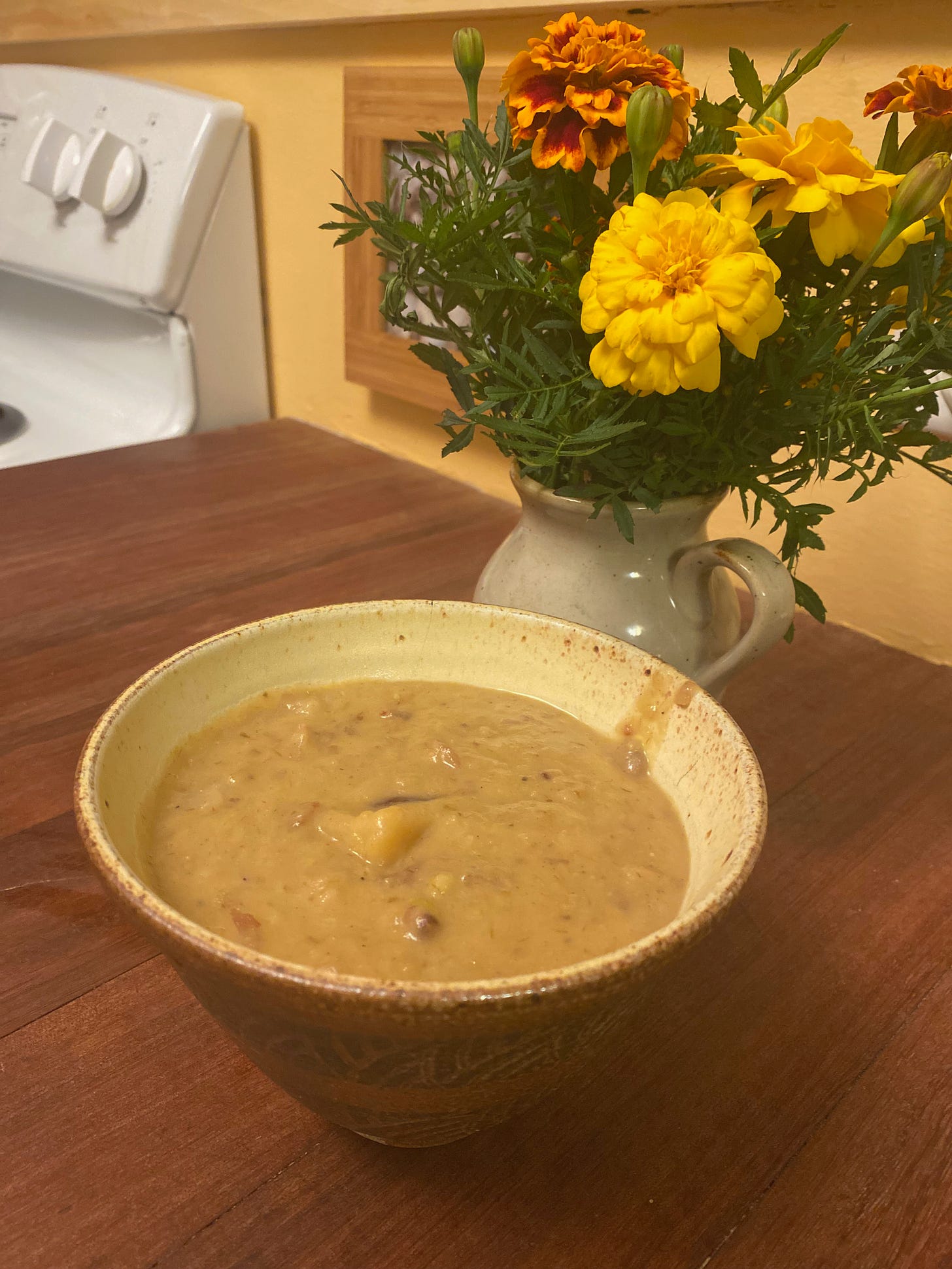 A ceramic bowl of chunky, creamy potato leek soup sits on a wooden counter in front of a vase of marigolds. Part of the stove is visible in the background.