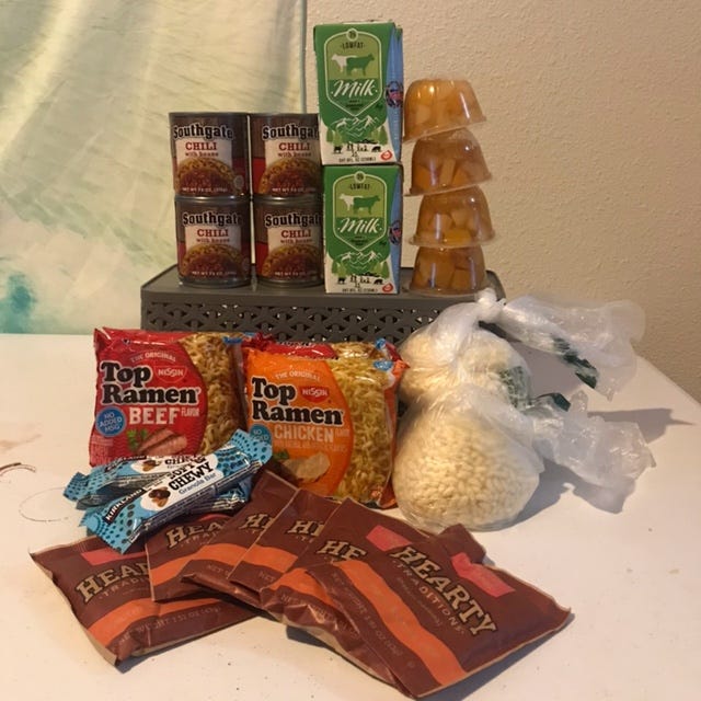A picture of what was inside the food bags: four cans of chili with beans, four individual cartons of milk, four mixed fruit cups, four packages of Top Ramen, six packets of Malt O'Meal, four soft and chewy chooclate granola bars, and two produce bags full of loose Rice Krispies and tied in a knot.