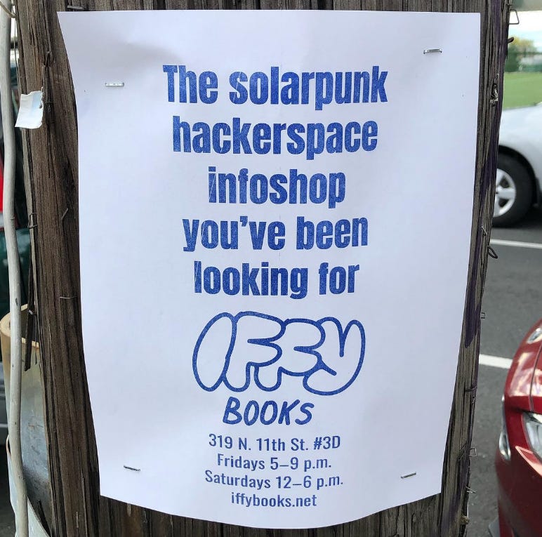 A sheet of A4 paper stapled to a telephone pole. On it is printed in blue: “the solarpunk hackerspace infoshop you’ve been looking for” and a stylized Iffy Books logo. 