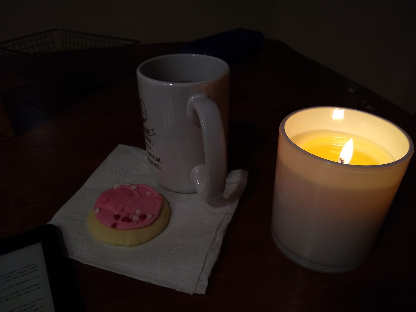 Corner of a Kindle, frosted sugar cookie on a napkin, mug, and a lit candle