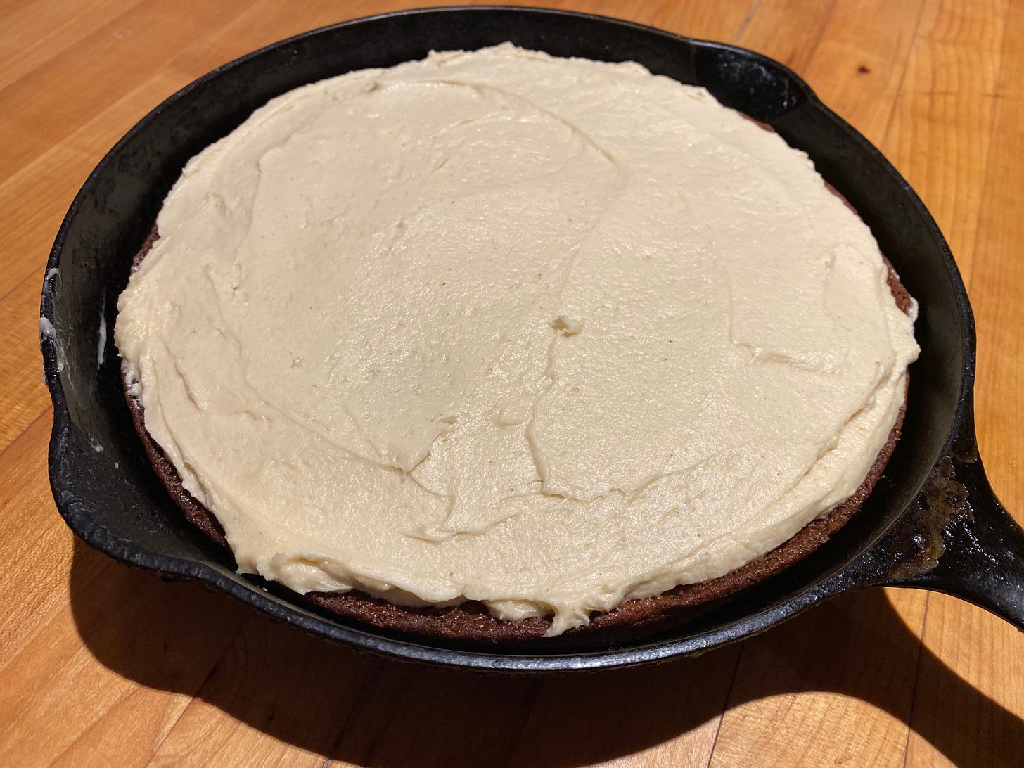 A round, white-frosted cake in a cast iron skillet sits on a wooden counter.