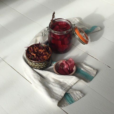 An open kilner jar filled with a pinkly red liquid and chunks of pickles. Beside it, a small bowl filled with orange and purple slaw, and a smaller bowl with turnip slices. These, encircled on one side by a white and green tea towel. 