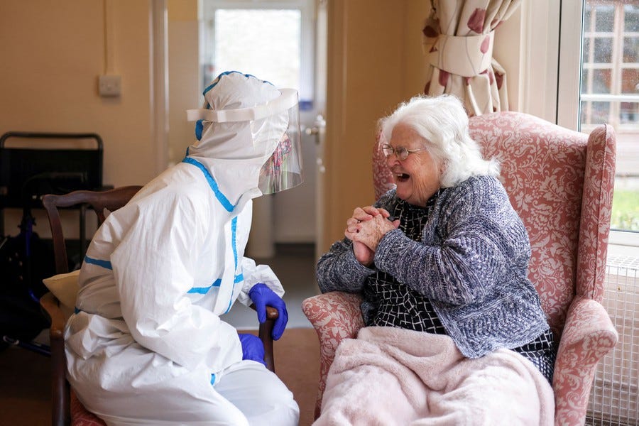 A woman in full protective gear sits in a chair beside her mother, who appears delighted.