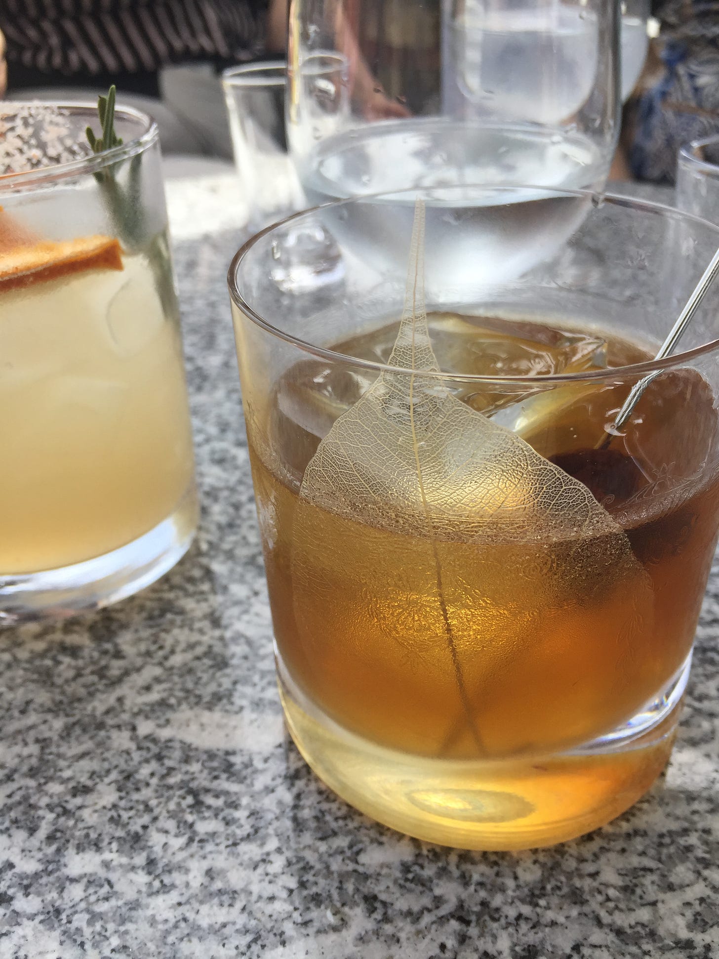 two glasses with cocktails in them on an outdoor table. In the foreground is an old fashioned with a skeleton leaf pressed into the side of the glass, and in the background is a paloma with a slice of dried grapefruit and a sprig of rosemary.