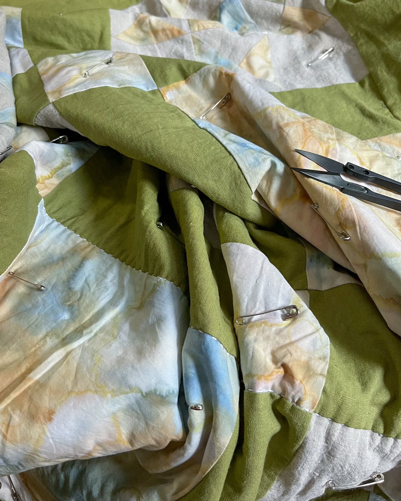 a basted quilt of green and tie dyed blocks with a pair of snips