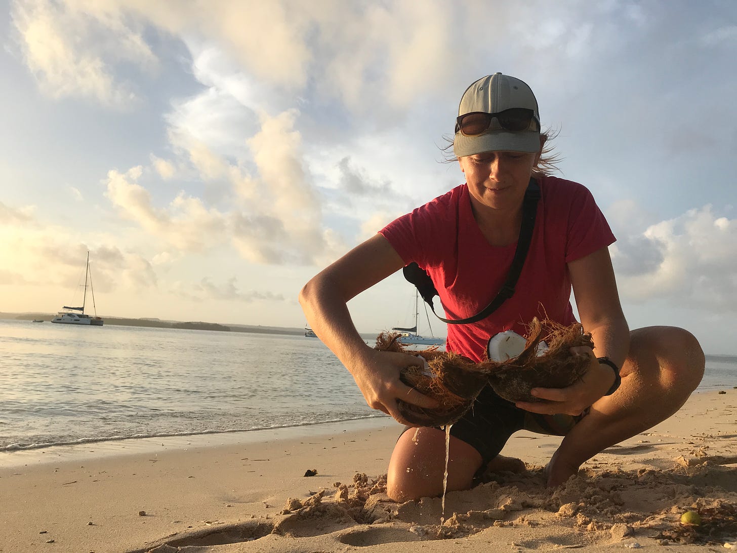 Trying to catch fresh coconut juice near Vava'u, Tonga.  Alt text: 30 year old woman on knees in shorts and t-shirt opens fresh coconut on sandy tropical beach with catamaran floating on sea in distance with puffy white clouds in blue sky above