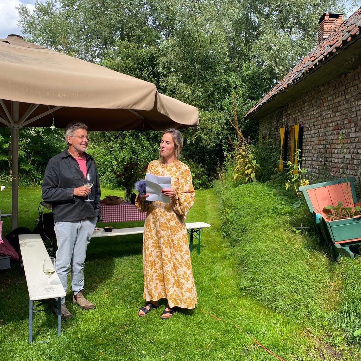 foto van Eva en haar vader die buiten in de zon in een tuin staa. Hij draagt een lichte broek en een donker vest en heeft een glas wijn vast. Zij heeft een gele bloemetjesjurk aan, een blaadje en een microfoon vast.