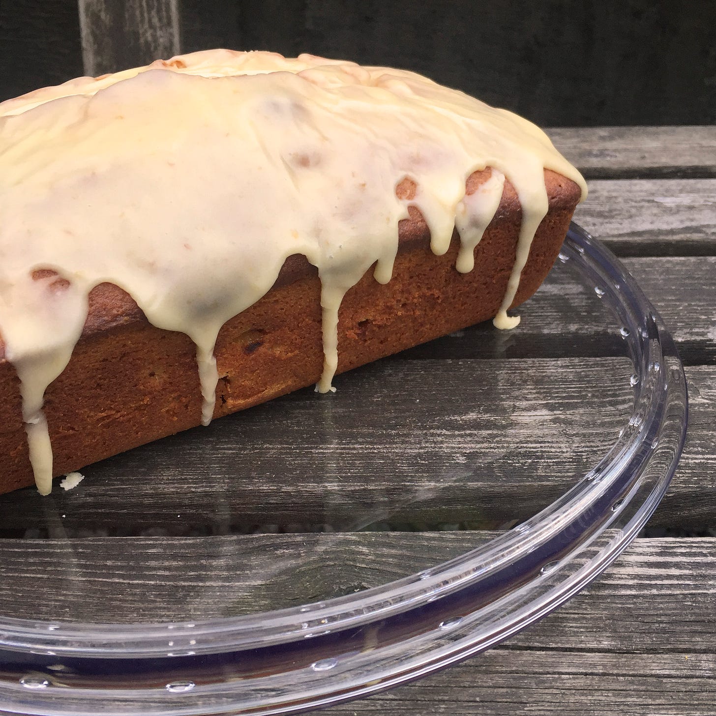 On a wooden bench sits a clear glass cake plate. On it is a loaf-shaped poundcake, lightly browned and covered in a peachy glaze that has dripped artfully down the sides.
