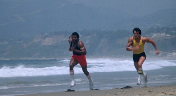 Rocky 3 still: Apollo Creed and Rocky run on the beach.