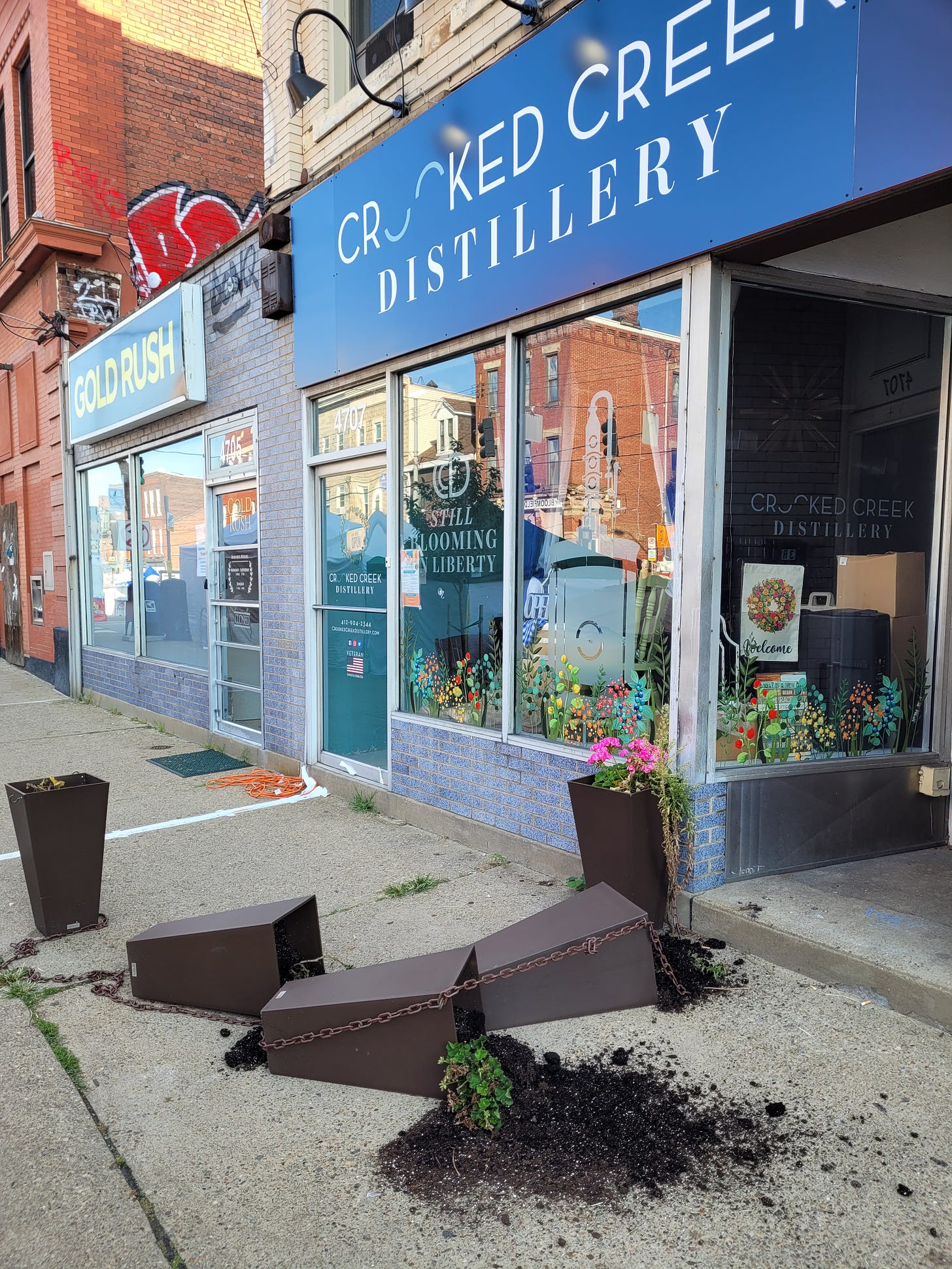 three overturned square planters litter dirt on the sidewalk in front of a shop called Crooked Creek Distillery