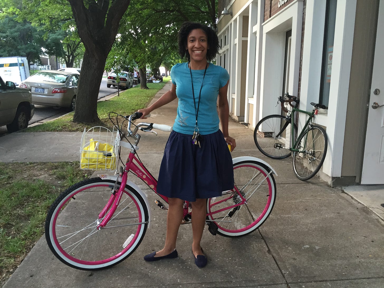 Kristen, in a blue outfit, stands in front of her pink bike on the sidewalk