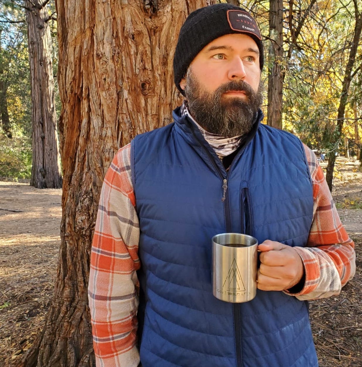 A bearded man in an orange flannel, blue vest, and grey wool stocking cap leans up against tree at a campground. He has a stainless steel coffee mug filled with coffee held about waist high.
