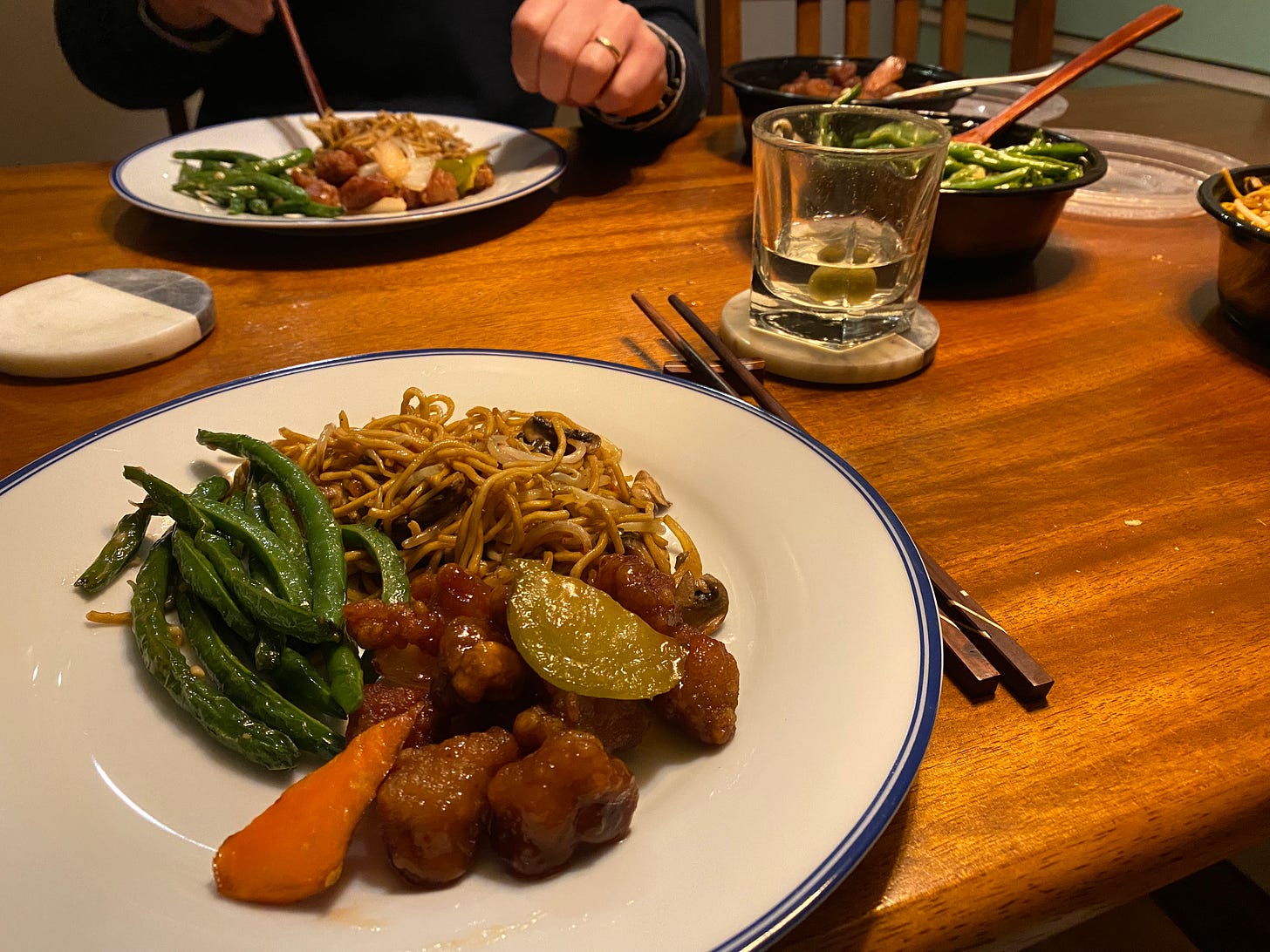 Two plates with Chinese food: green beans in garlic sauce, mushroom chow mein with thin noodles, and sweet and sour pork with carrots and peppers. The takeout containers are visible at the right corner of the frame. Chopsticks sit next to the plate in the foreground.
