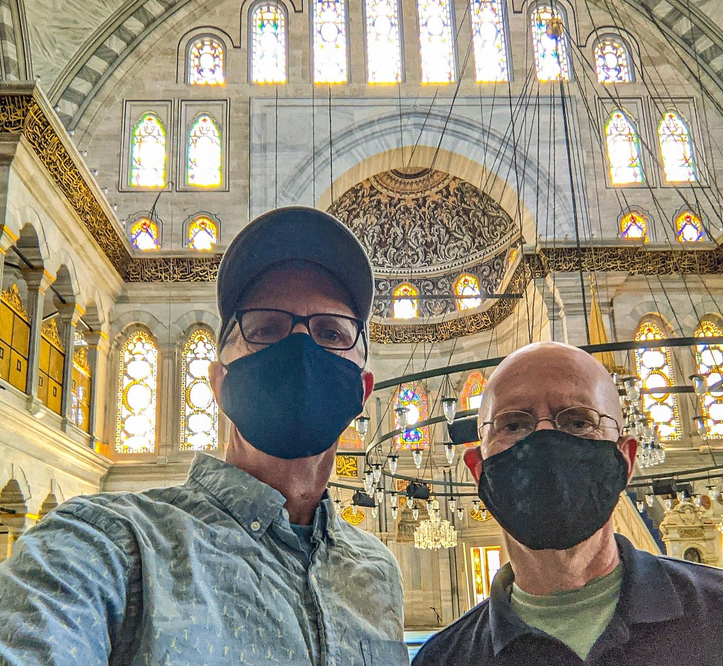 Brent and Michael wearing masks and visiting a mosque in Istanbul. The mosque is made of white marble with stained glass windows and golden calligraphy against a black background.
