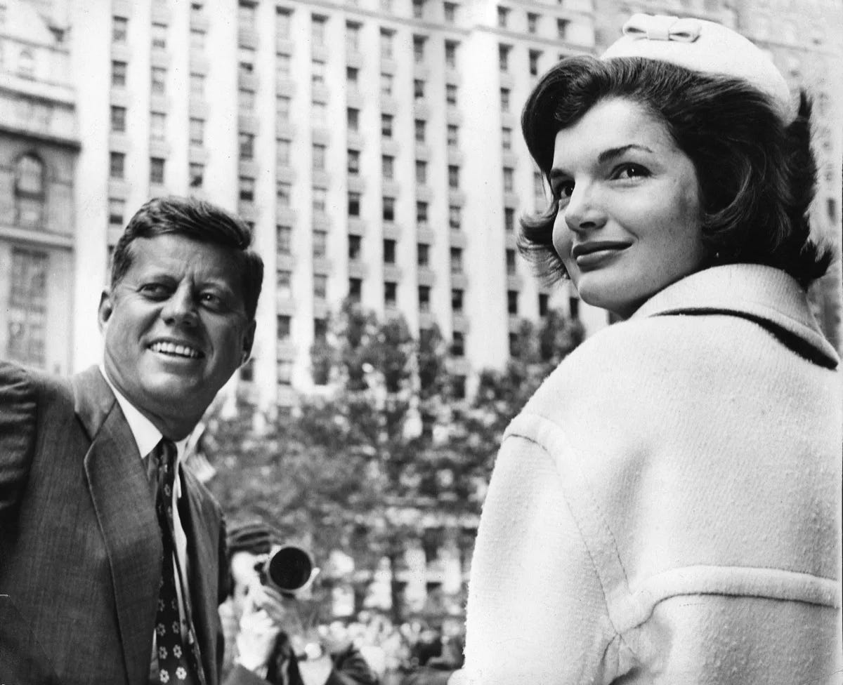 This black-and-white image shows John F. Kennedy on the left and Jacqueline Bouvier Kennedy of the right. Both look behind them and to the left, smiling at something off-camera. Visible in the distance is a man shooting their photo with a camera and, behind him, a large, white, high-rise building. This black-and-white image shows John F. Kennedy on the left and Jacqueline Bouvier Kennedy of the right. Both look behind them and to the left, smiling at something off-camera. Visible in the distance is a man shooting their photo with a camera and, behind him, a large, white, high-rise building.
