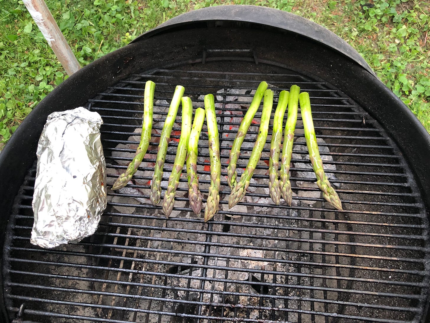 Asparagus grilling on a hot charcoal grill. Beside them on the grill is a foil packet.