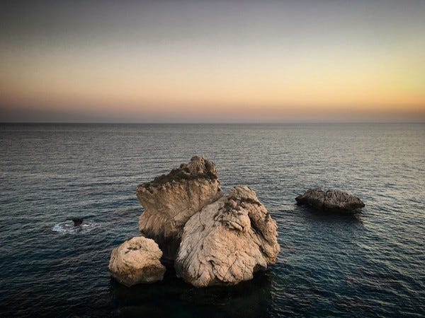 Rochedos no litoral de Pafos, no Chipre. Foto de Sergei Gussev/Flickr. Descrição de Imagem: Em um mar vasto e calmo, um grande aglomerado de três pedras grandes e cor de areia está próximo da foto. Na direita, um rochedo menor e mais ao fundo. O céu está sem nuvens.