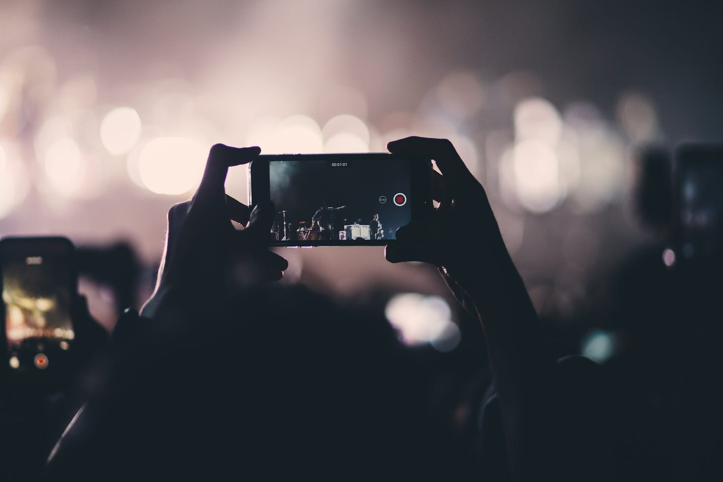 Photo of someone holding a mobile phone in landscape above a crowd, videoing a performance on the stage in the distance