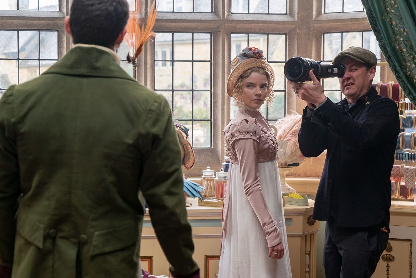 Photo shows actress Anya Taylor-Joy in Regency gown, surrounded by a cameraman on the set of the 2020 film 'EMMA'. Approaching is the character of Frank Churchill, shown from the back. 