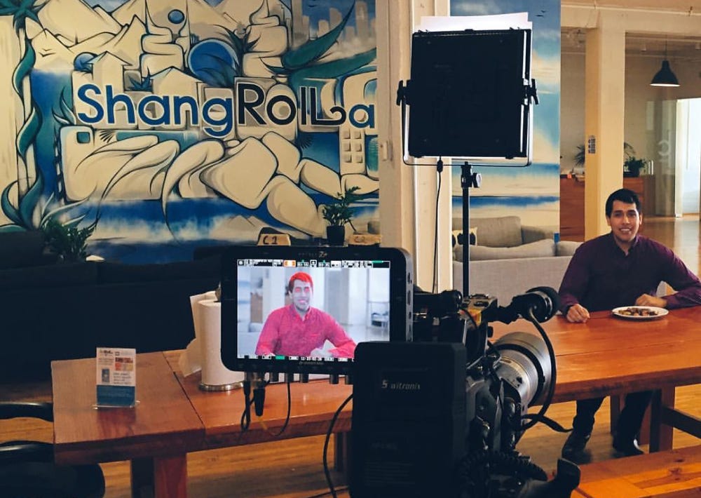 Photo of Kevin in the background sitting at a table recording a marketing video with the ShangRoll mural in the background.