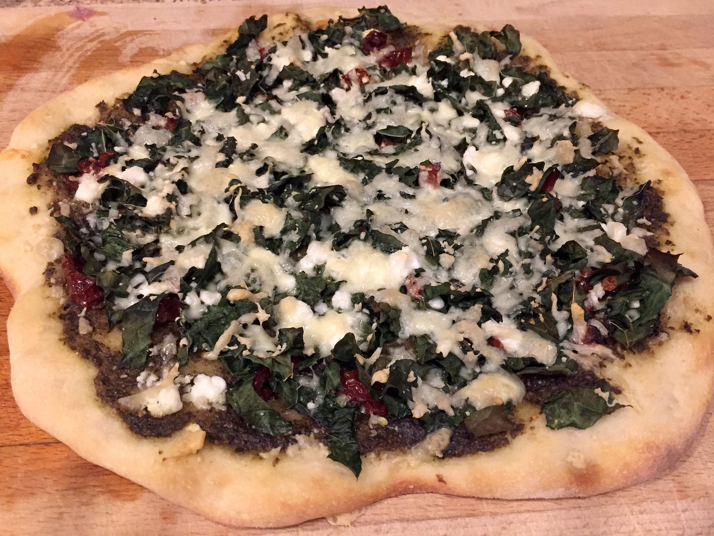on a wooden cutting board, a hand-stretched, uneven pizza with a pesto base, kale, and sundried tomatoes. Crumbles of feta and melted, lightly browned mozza sit on top.