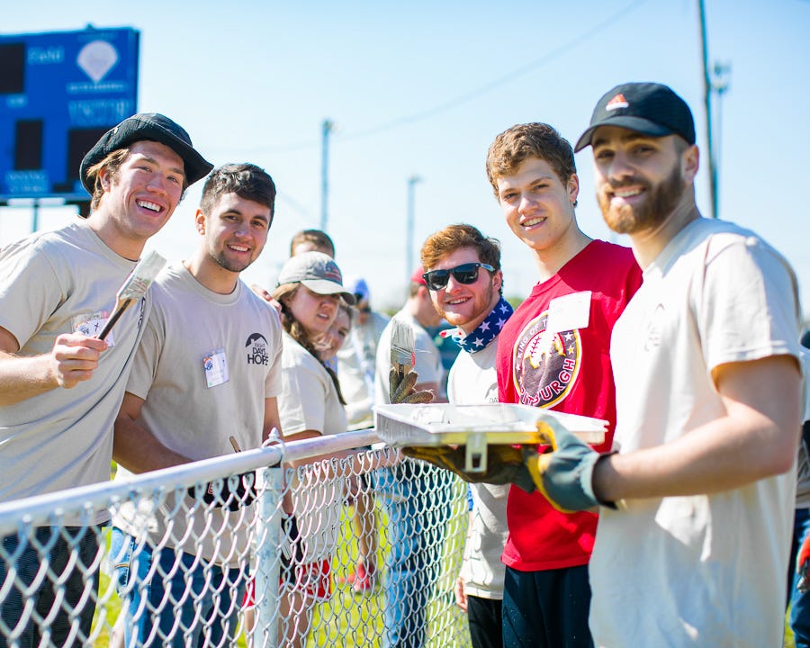 Teenage boys and girls painting a chain link fence near an athletic field.