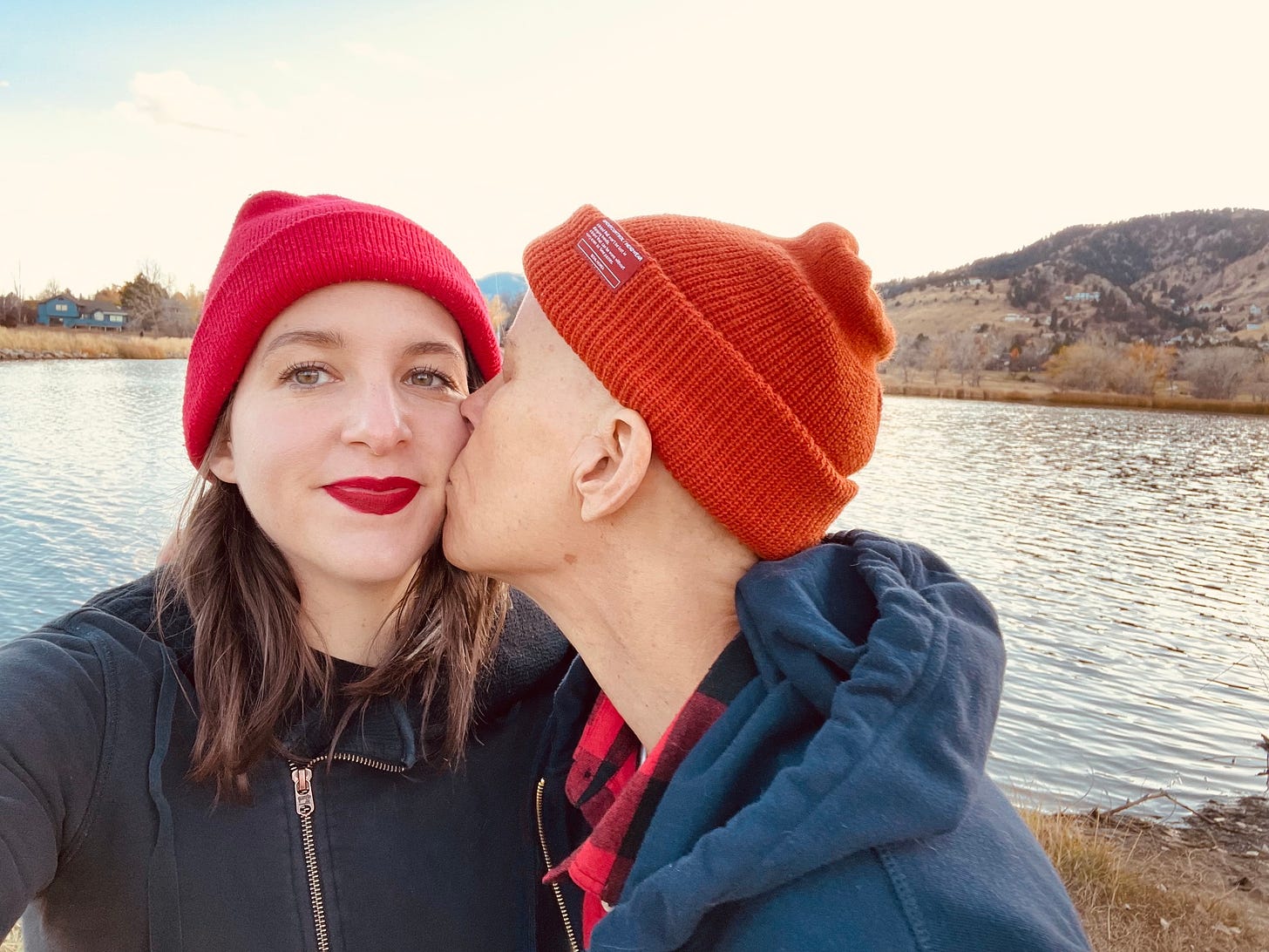 Andrea’s partner Meg wears a red beanie and red lipstick. Andrea, in an orange beanie, is kissing her on the cheek. In the background is a lake with mountains behind them.