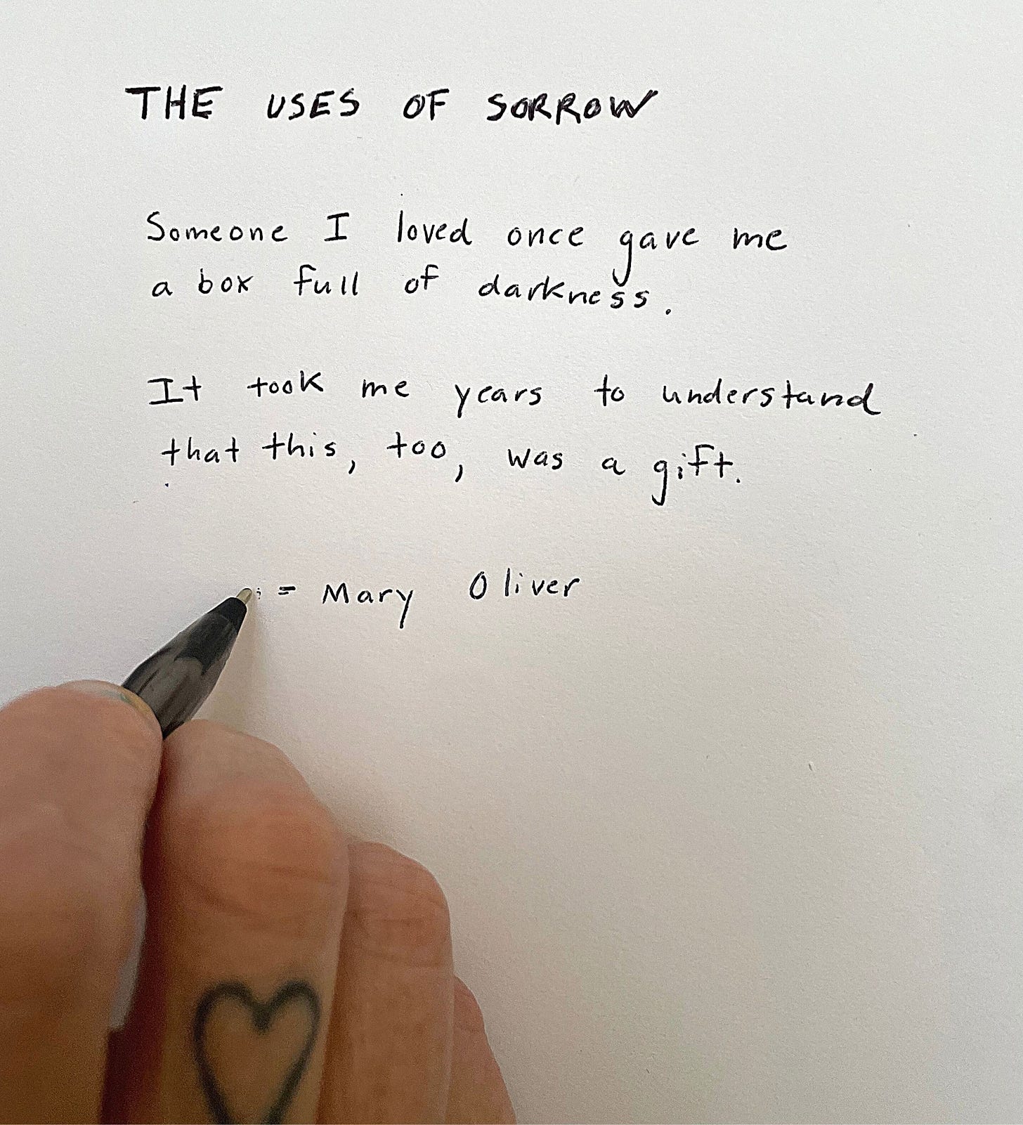 Andrea’s hand with a heart tattoo above their middle finger knuckle and pen in hand writing a quote. The quote reads: “ THE USES OF SORROW Someone I loved once gave me a box full of darkness. It took me years to understand that this, too, was a gift. - Mary Oliver”