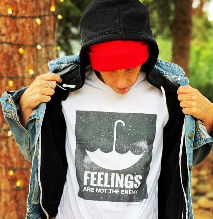 A photo of Andrea Gibson with a black and white t-shirt that says “Feelings are not the enemy” with an upside down umbrella graphic. They are also wearing a jean jacket, black hoodie and red beanie. In the background is a tree circled with fairy lights. 