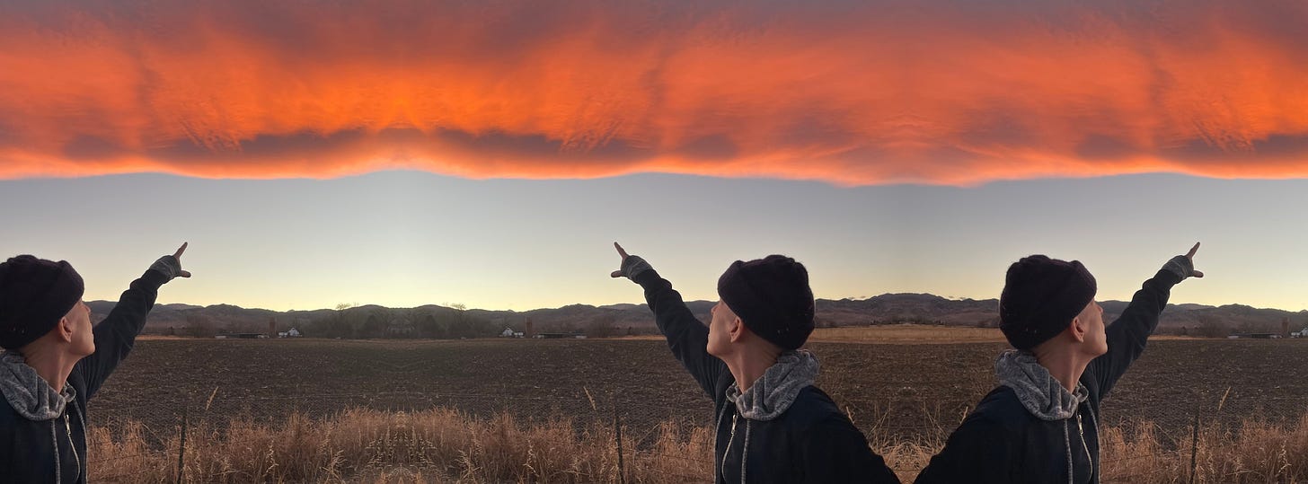 A mirrored photo of Andrea Gibson in a field pointing at a fiery orange cloud in the distance. Because of the mirroring effect, it looks like there are 3 Andrea’s pointing.