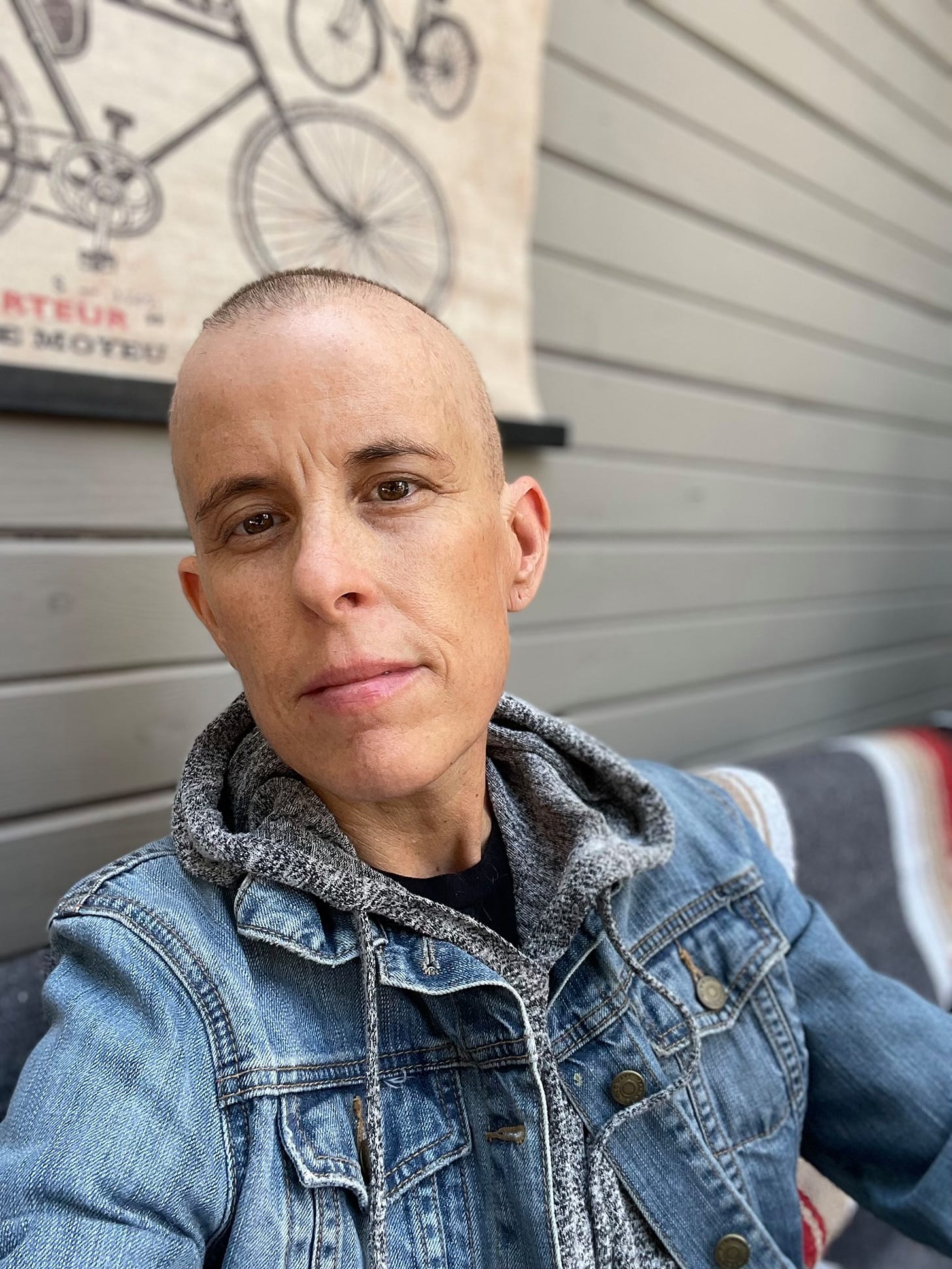 Selfie of Andrea Gibson with very little hair, and full eyebrows. They are wearing a grey hoodie with a jean jacket over it. 