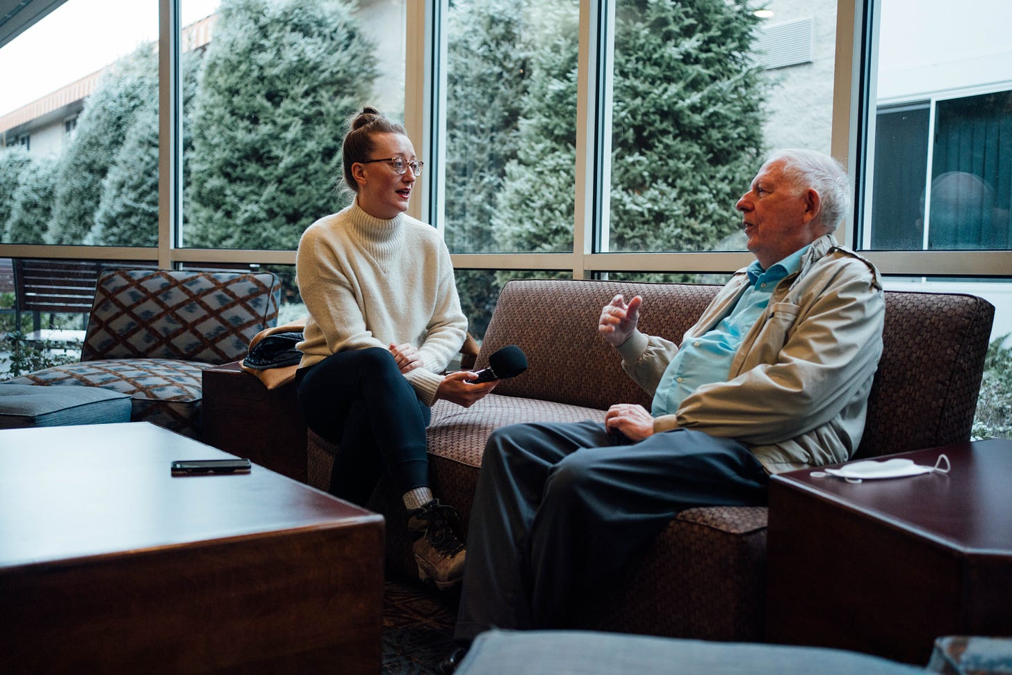 Garnet and Tony sit on a couch speaking. Garnet is holding a recorder in one hand and Tony is gesturing with one of his hands. There are trees visible out the window with a light dusting of snow.