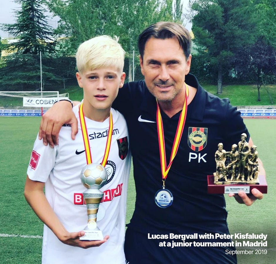 A photo featuring a young Lucas Bergvall with his former junior coach, Peter Kisfaludy. Kisfaludy has his arm over Bergvall's shoulder and both are looking into the camera while holding small trophies. In the background is a goal frame and pitchside trees.