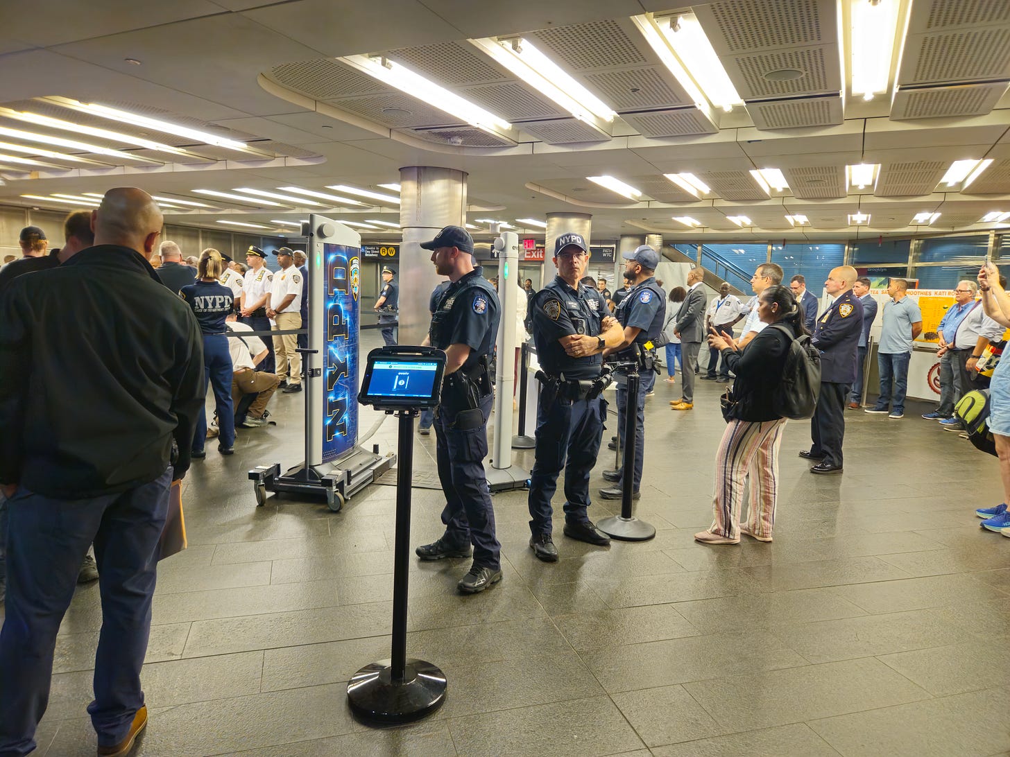 Photo of the Evolv system set up in the Fulton St. Subway Station as the start of the pilot program, and people watching the press conference announcing the pilot.