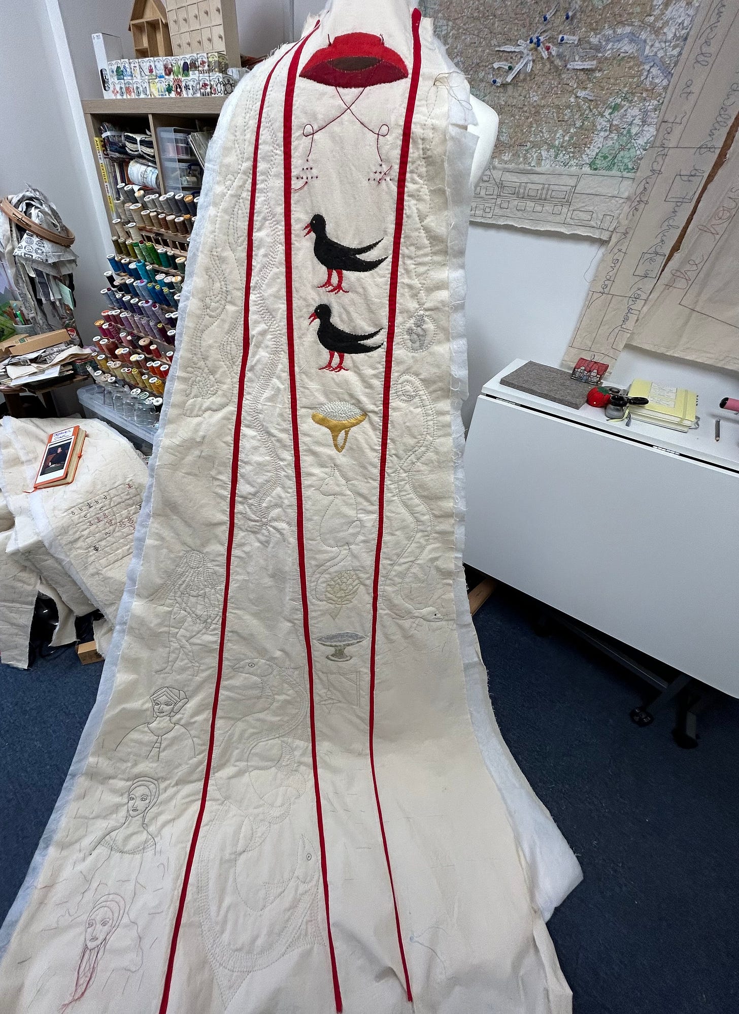 A stitched work in progress hanging on a tailor’s mannequin. The work is in cream calico with motifs stitched and painted - a cardinal’s hat, two black birds, a ring, a cat, an artichoke, an eel, a comet, fish, a snake and other motifs. It is situated within an artist’s studio, with thread and notebooks visible, and various reference materials.