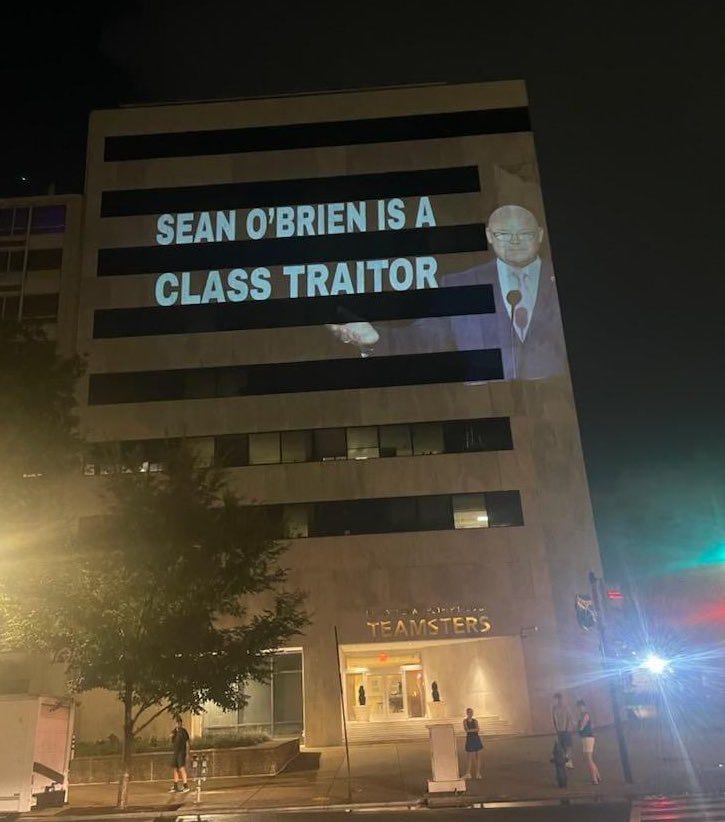 The words “sean O’Brien is a class traitor” projected onto a blocky tan building (the teamsters hq in DC)