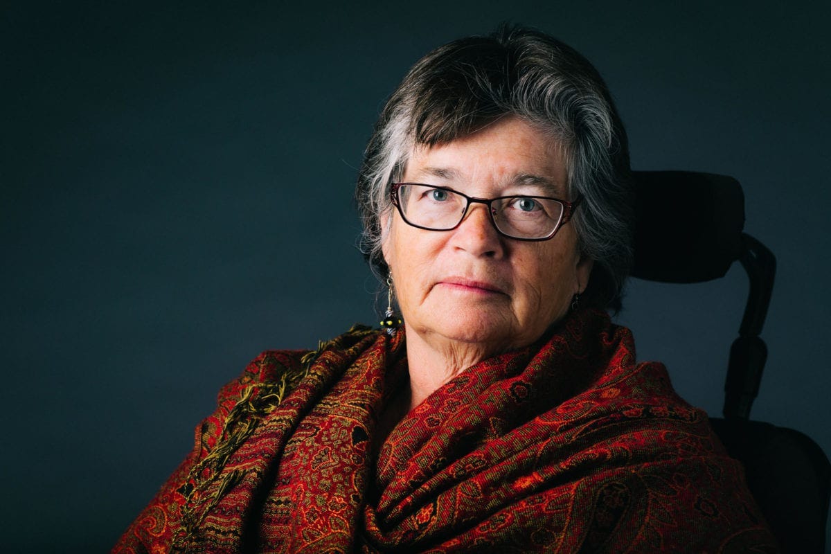 A white woman with gray and black and hair, wearing glasses and a colorful scarf, sits in a wheelchair in front of a neutral background. 