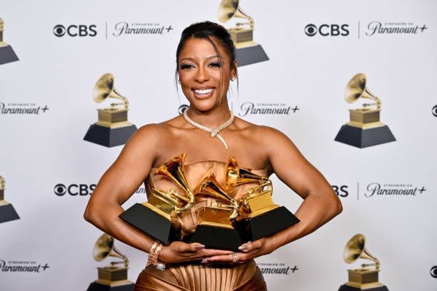 Picture of singer Victoria Monét wearing a strapless brown gown holding three Grammy awards.