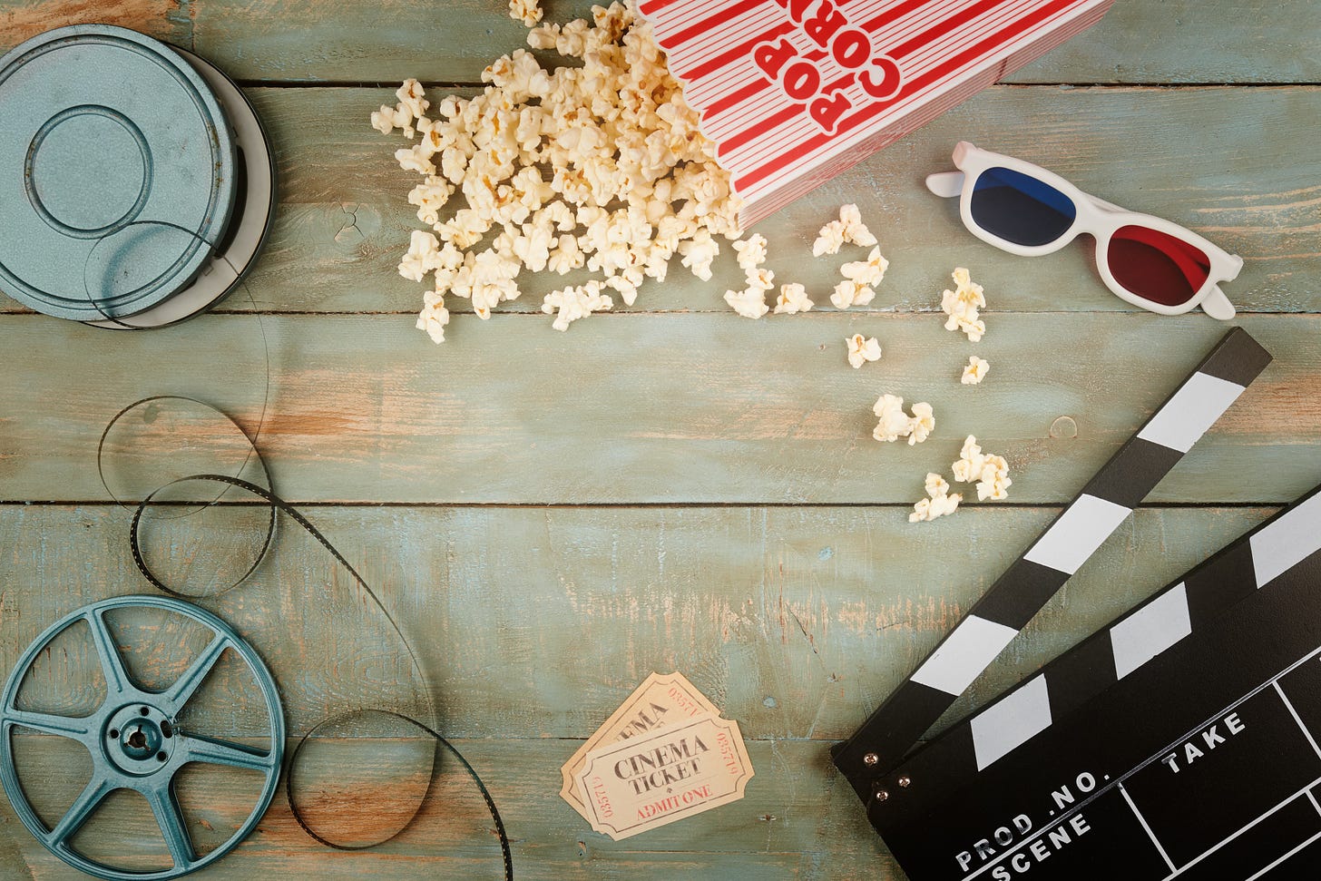 Retro cinema objects on wooden background. A metal film reel case and the reel, with the film strip curled between them; two old-fashioned cinema tickets; a black and white movie clapper, white-framed 3D movie viewing glasses, and a popcorn bucket laying flat, spilling popcorn over the background.