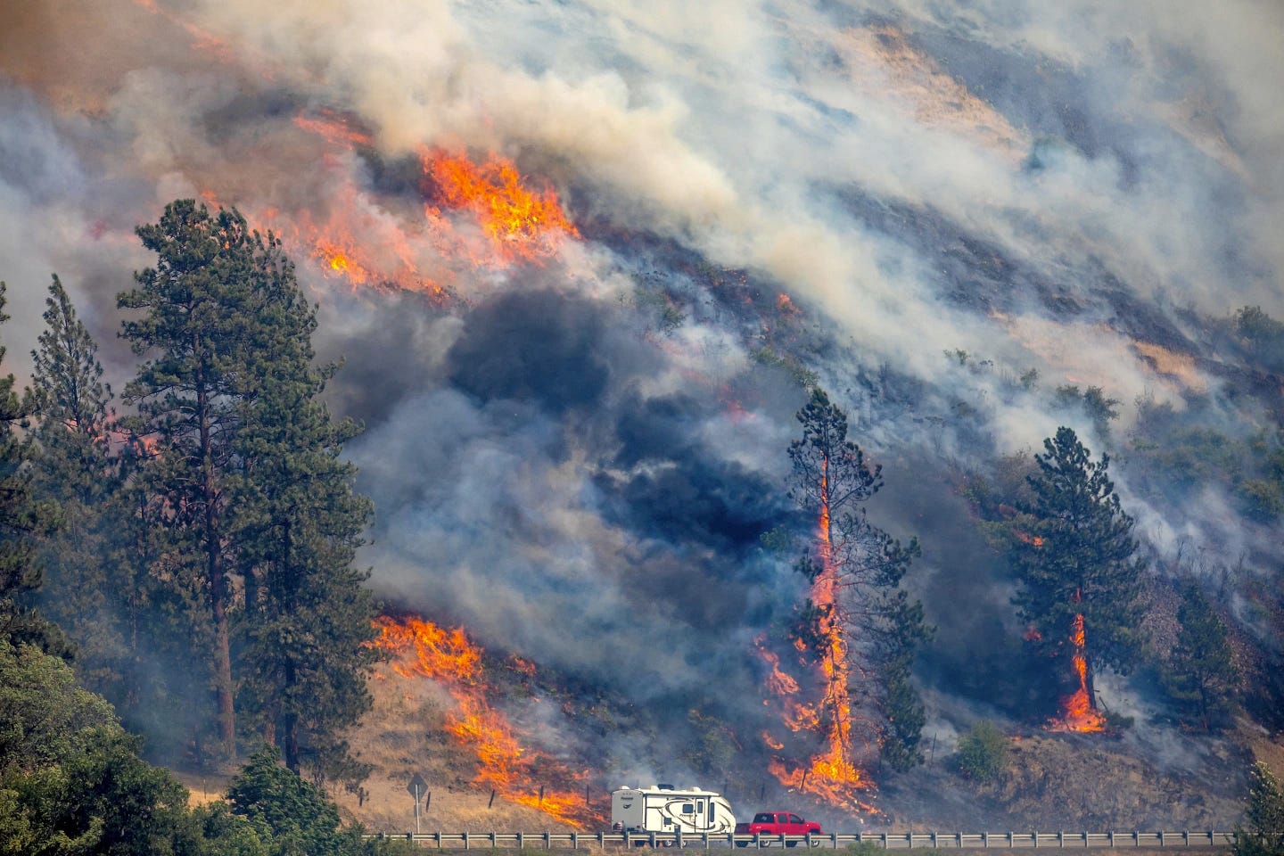 Red pickup truck pulling camper drives by a mountain on fire in Myrtle, Idaho.