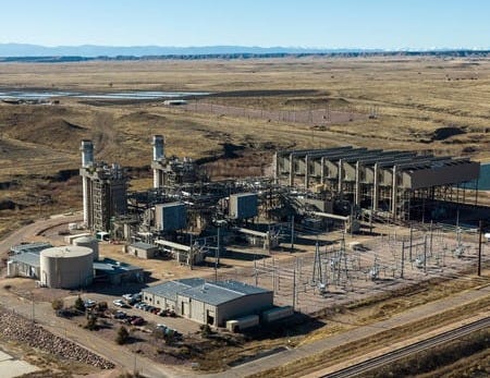 Aerial image of Front Range Power Plant. It features two smokestacks, which are not very prominent. There are overhead lines and substation equipment next to the plant and a vast array of industrial equipment next to the stacks. The surrounding landscape is brown grass.