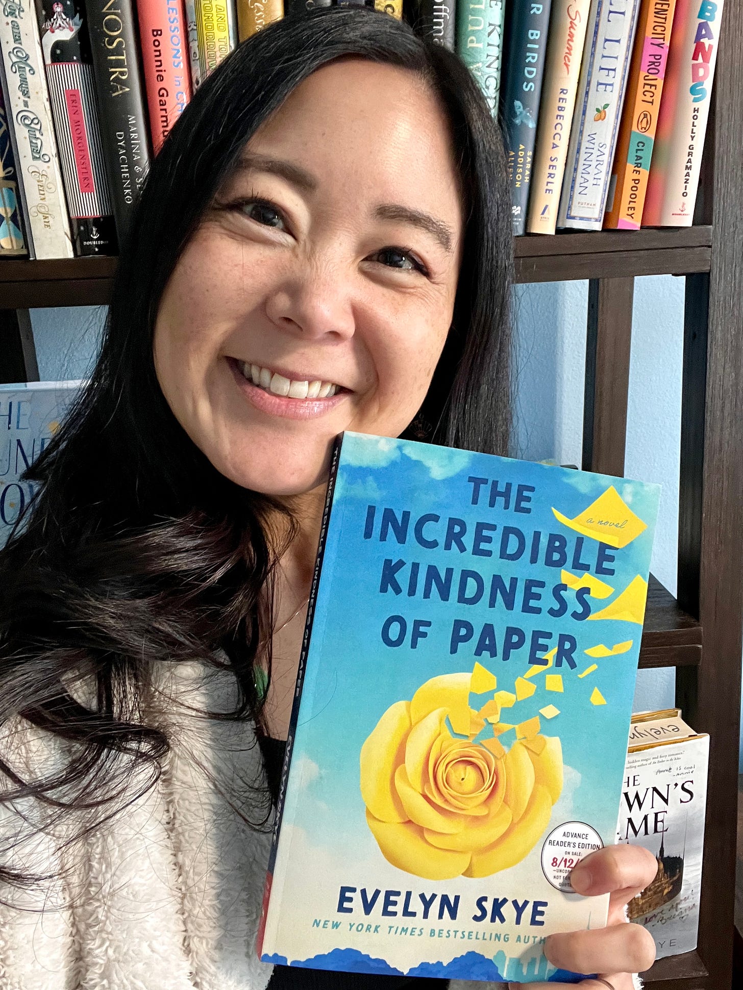 Author Evelyn Skye in front of a bookcase, holding the ARC of her next novel, The Incredible Kindness of Paper