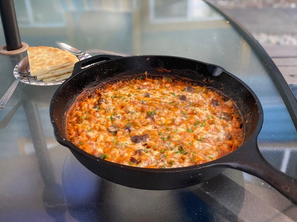 A cast iron pan filled with white beans in a deep orange tomato sauce and covered in melted cheese. Chopped olives are visible throughout. On a small plate in the background are triangles of naan and two spoons.