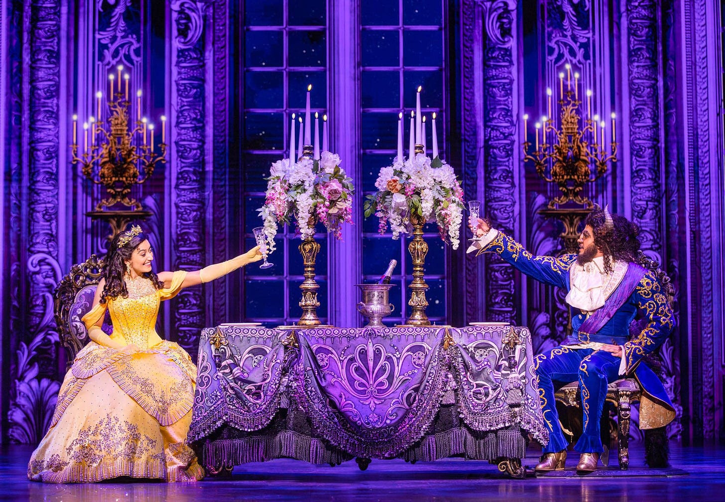 A photograph from onstage of the musical "Beauty and the Beast". It shows the characters Belle and the Beast sitting at dinner together, raising their glasses in a toast. They are sat in a magnificent castle bathed in purple light. Belle is wearing a beautiful yellow ball gown and tiara. The Beast is dressed in a blue suit with gold accents.